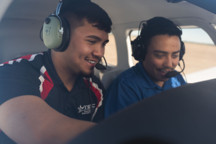 Two people sit in the cockpit of a small airplane. They're wearing headsets and leaning in towards a console, smiling.
