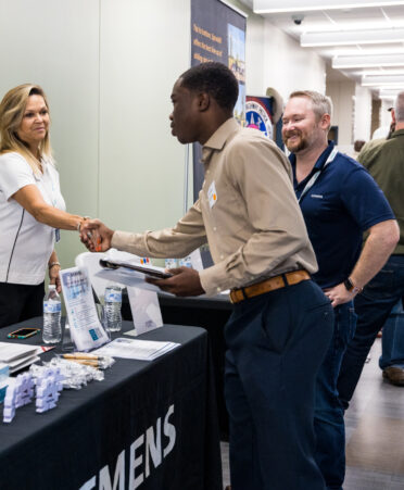 A student shakes hands with a company representative over a table containing company information.