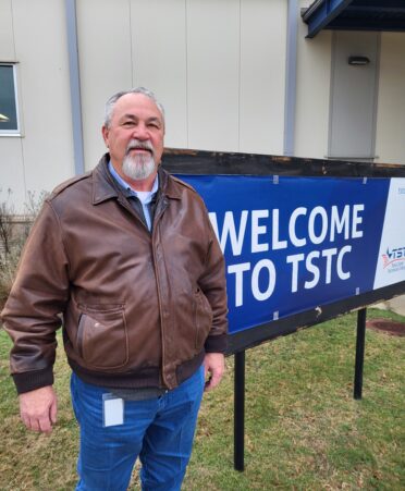 Steve Abernathy, wearing a brown jacket, stands in front of a sign reading welcome to TSTC.