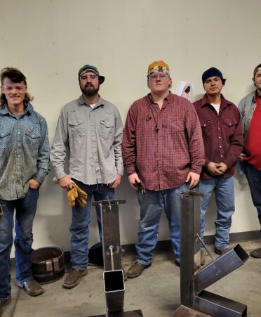 TSTC students stand against a wall with welding projects in front of them.