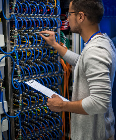 Back view portrait of young man connecting wires in server cabinet while working with supercomputer in data center