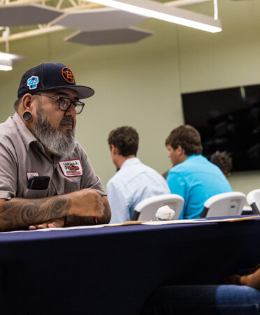 A student sits across a table from a coach wearing a hat. Two students sit in the background.