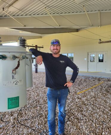 Brandon, wearing a navy blue TSTC shirt and jeans, leans against a transformer used for teaching.