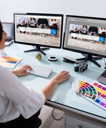 A woman wearing a white shirt sits at a desk working on two screens.