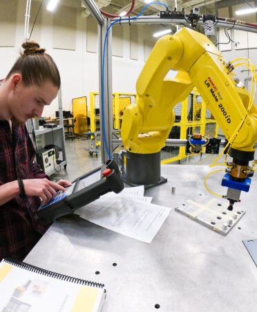 A student wearing a plaid shirt looks at a device that controls a yellow robotic arm