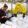 A student wearing a plaid shirt looks at a device that controls a yellow robotic arm