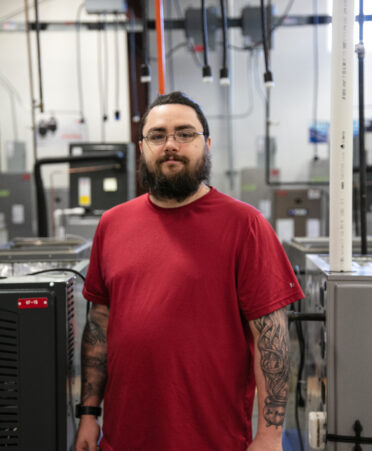 HVAC student stands between training units wearing a red t-shirt and glasses. He has long dark hair and a dark beard. He has tattoos on both arms.