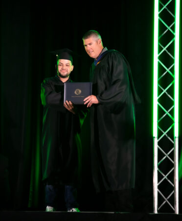 A student stands on the stage at graduation, shaking hands and taking a photo with Marcus Balch, provost of the North Texas campus. They hold a diploma between them as they pose for a photo.