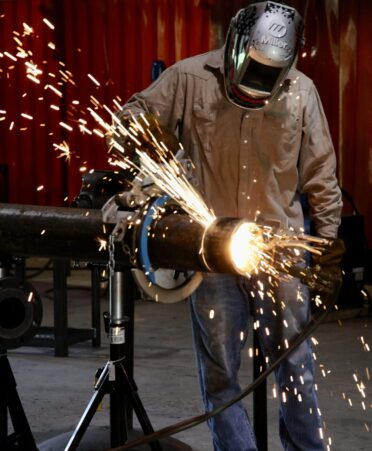 A welding student in a tan shirt and welding mask cuts a pipe to the appropriate length