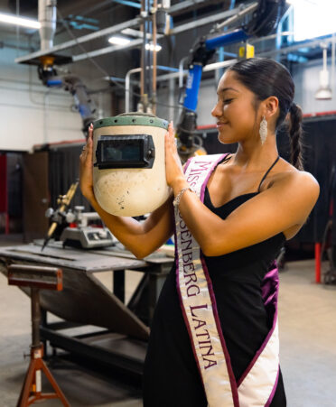 Mendez, wearing a dress and the Miss Rosenberg Latina ribbon holds a welding helmet to her side