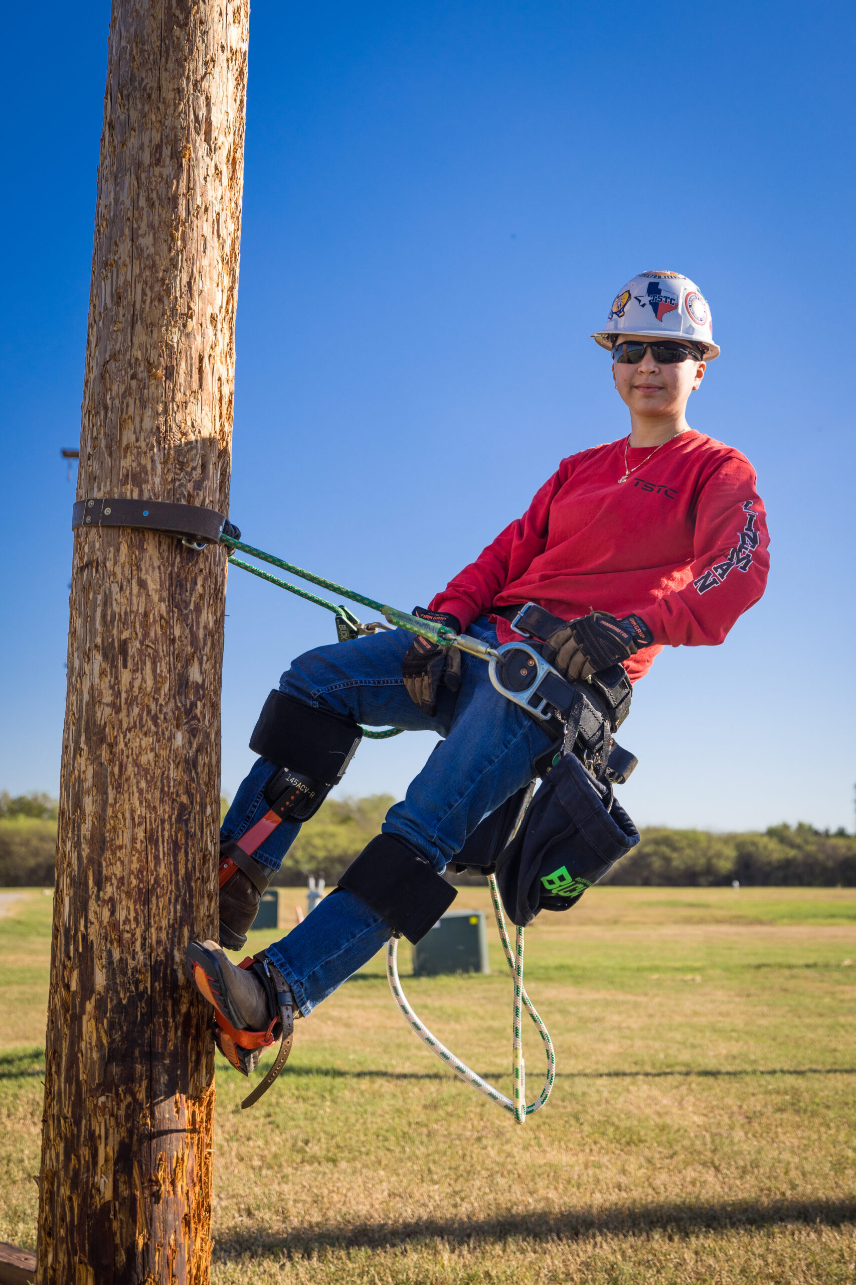 Kelly wearing jeans, a red shirt and safety gear, stands hanging on a harness.