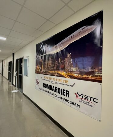 Shows Bombardier banner, with a large jet flying over a city. There is white space below that has the Bombardier and TSTC logos as well as the words aviation apprenticeship program below the Bombardier logo. The banner is on a wall in a long hallway of classrooms.