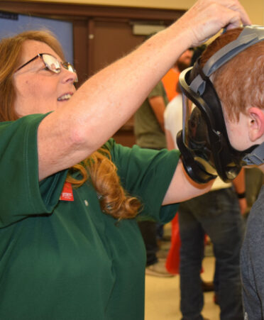 A woman in a green shirt putting a gas mask on a teenager