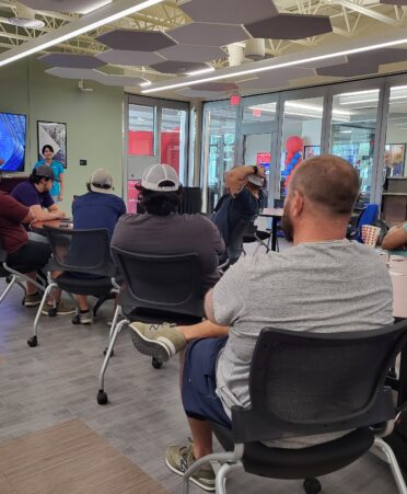 Students sit around tables in a conference room and look toward representative giving presentation on tv