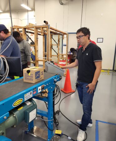 Nguyen stands in a black shirt and jeans operating a conveyor belt with a box on it.