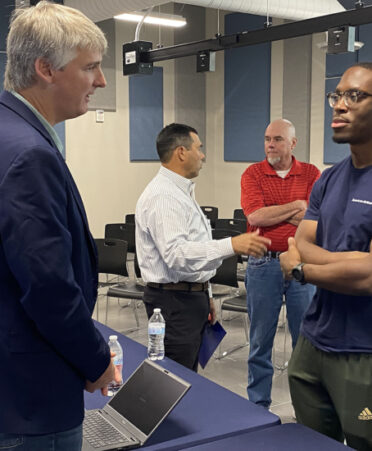 Man in sports jacket talking to man in blue tshirt