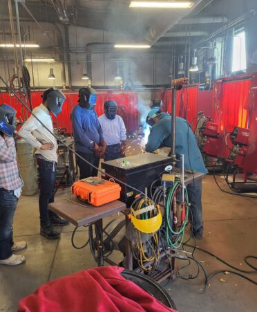 Four students wearing welding helmets gather around an instructor in a blue shirt welding. Equipment lies in front of the group