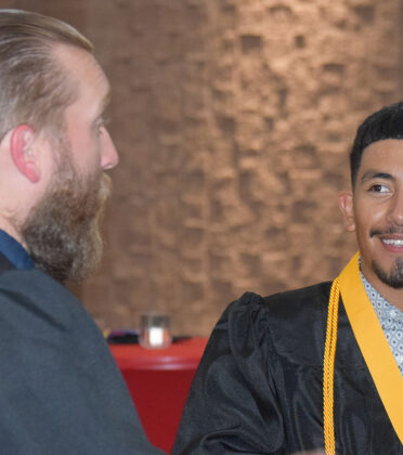 a bearded man talks to a man with a graduation gown and cord