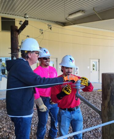 Teacher wearing a blue sweater and white hardhat teaches two students wearing a red shirt and pink shirt respectively. Pole and wires in the foreground.