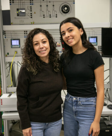 Mother and daughter stand side by side in front of one of the electrical training boards, smiling at the camera. Both wear blue jeans and black tops.