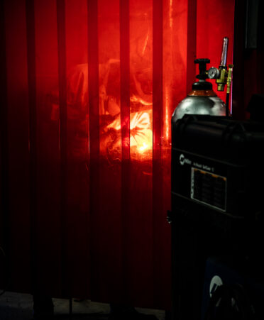view of student with welding helmet down, welding. The photo is taken through a red protective curtain.