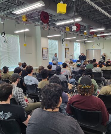 Students sit in rows while presenters talk at the front of the group