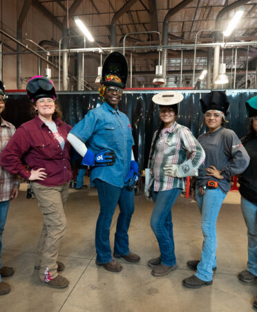 Six female welding students stand in welding shop in a line with gear on, smiling at the camera