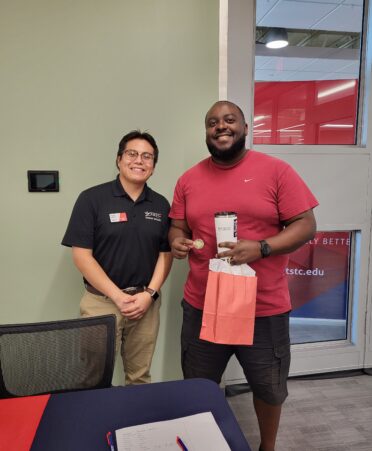 Ruben and Kelvin stand next to each other, in a black and red shirt respectively. Kelvin holds gifts given to him.