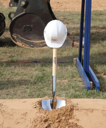 A TSTC branded shovel sits in dirt with a white hard hat resting on top of it.