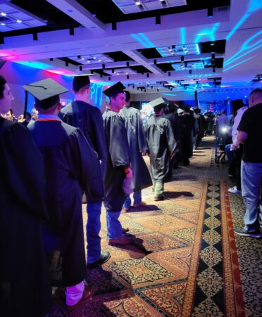 Students wearing graduation gowns and caps walk away from camera while family watches from right side.