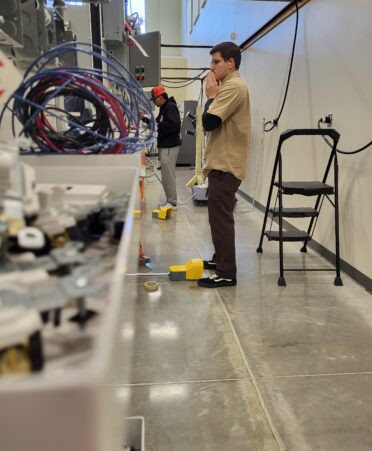 A student stands with his hands clasped together looking at some equipment. Wires and bolts sit in the foreground.