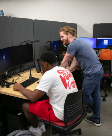 Student sits at desk with monitor while instructor stands beside him, leaning over, offering assistance.