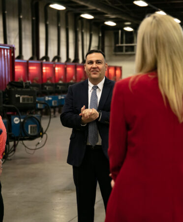 Joe Esparza stands in front of line of welding booths in a suit and tie, smiling with hands clasped while talking with a welding instructor and two others.