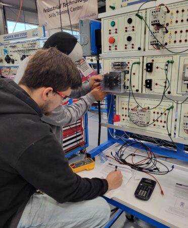 Two students, one in a dark hoodie and one wearing a ball cap work on some equipment, paper, and wires.