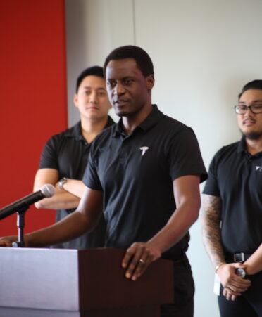 Anthony stands at a podium wearing a Tesla shirt with his instructors in the background