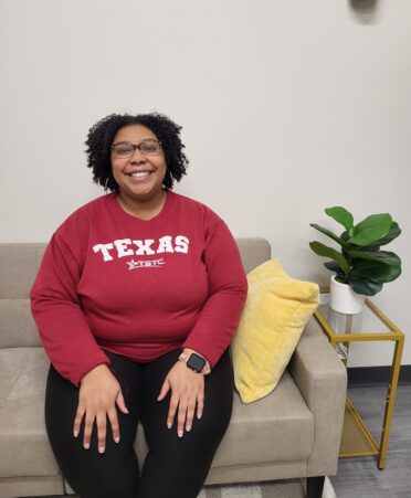 Justice, wearing a red TSTC sweater sits on a beige couch with a plant and pillow next to her.