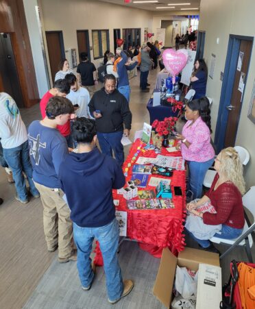 Students form a half-circle around a booth that has goodies and information on it. Representatives stand ready to help them.