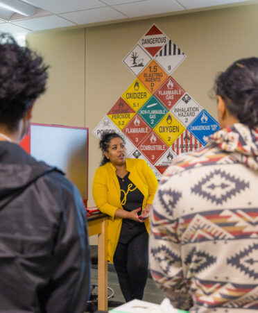 Andrea leans on a table wearing a yellow blazer and black shirt. Students wearing a black jacket and a white and brown jacket face away from the camera toward Andrea