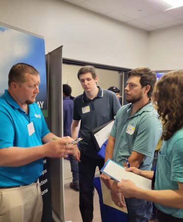 A industry partner representative wearing light blue meets with students who are wearing various shades of green.