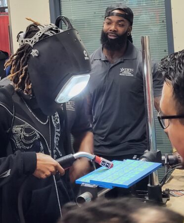 A student wearing a black hoodie and a simulation welding helmet holds a welding tool onto a blue panel.