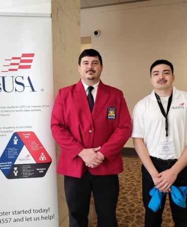 Austin wears a red jacket and Sergio stands on the right wearing a white shirt. A SkillsUSA sign stands next to them.