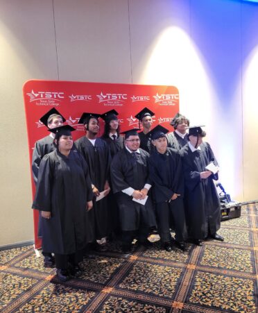 Students wearing graduation cap and gown stand for a picture with a tstc background