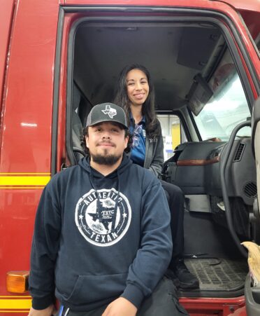 Andres leans against a red Diesel truck wearing a blue tstc hoodie and hat. Stephanie sits inside the truck behind him wearing a gray sweater over a blue shirt