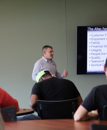 Students wearing black, red and gray shirts sit facing away from the camera. In the center in Michael Wilkinson presenting to the students.