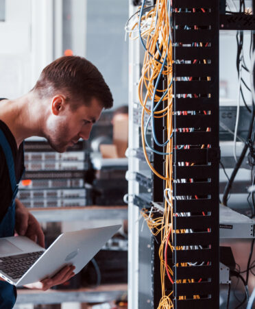 Young man in uniform and with laptop works with internet equipment and wires in server room.