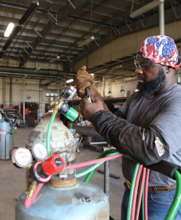 James Norman, wearing jeans, a gray shirt, and a red and blue welding cap works on some metal canisters