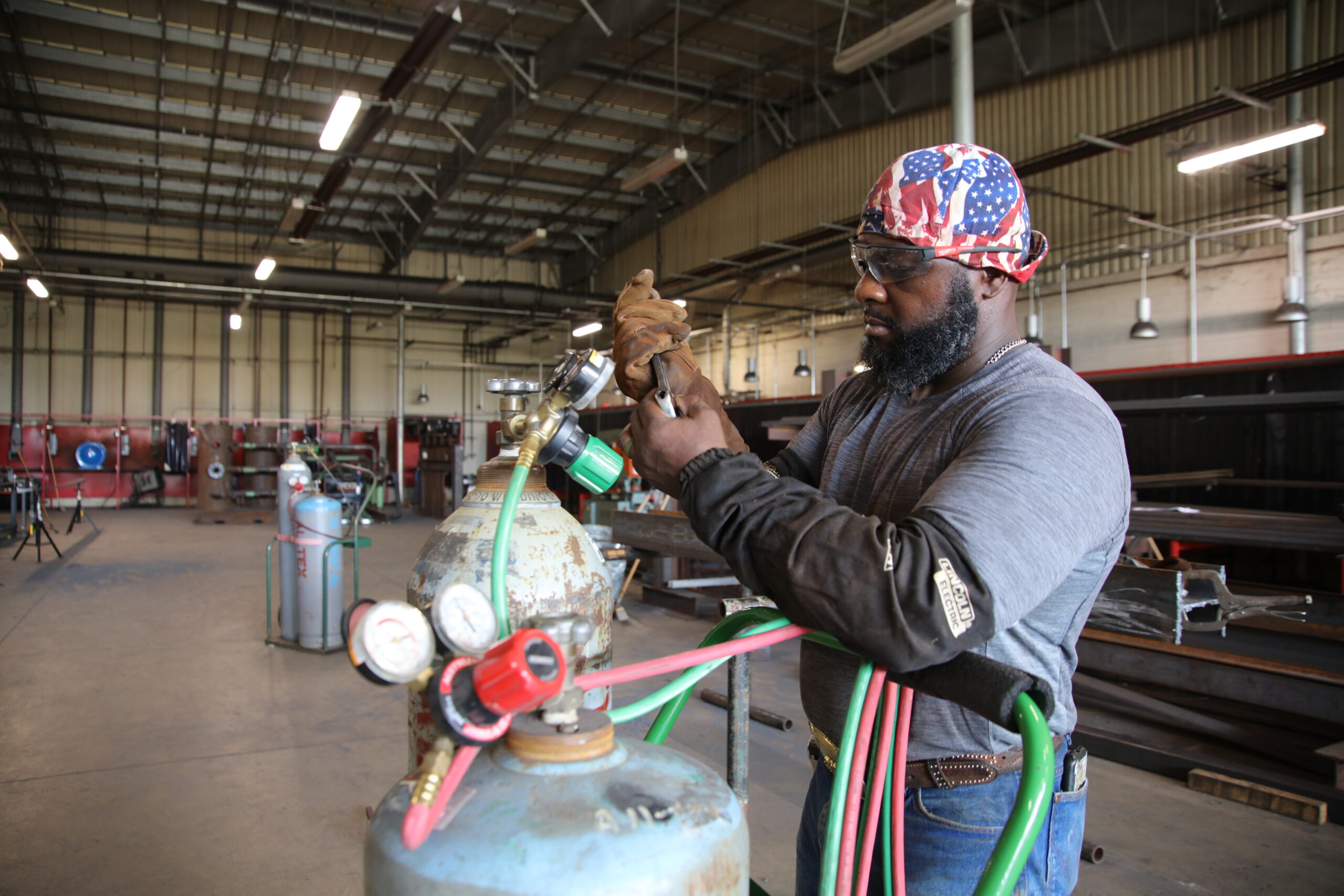 James Norman, wearing jeans, a gray shirt, and a red and blue welding cap works on some metal canisters