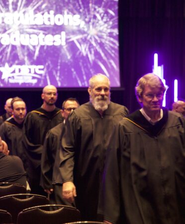 Students and faculty in graduation attire exit a commencement ceremony. Behind them a large screen displays the text "Congratulations graduates" with white fireworks.