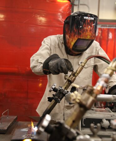 Eric, wearing gloves, a long sleeve shirt and a flame decorated welding helmet works with welding tools.
