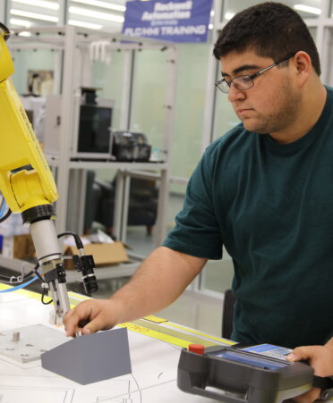 Delgado works with a yellow robot. He wears a dark green shirt and glasses and is holding a large remote for the robot.